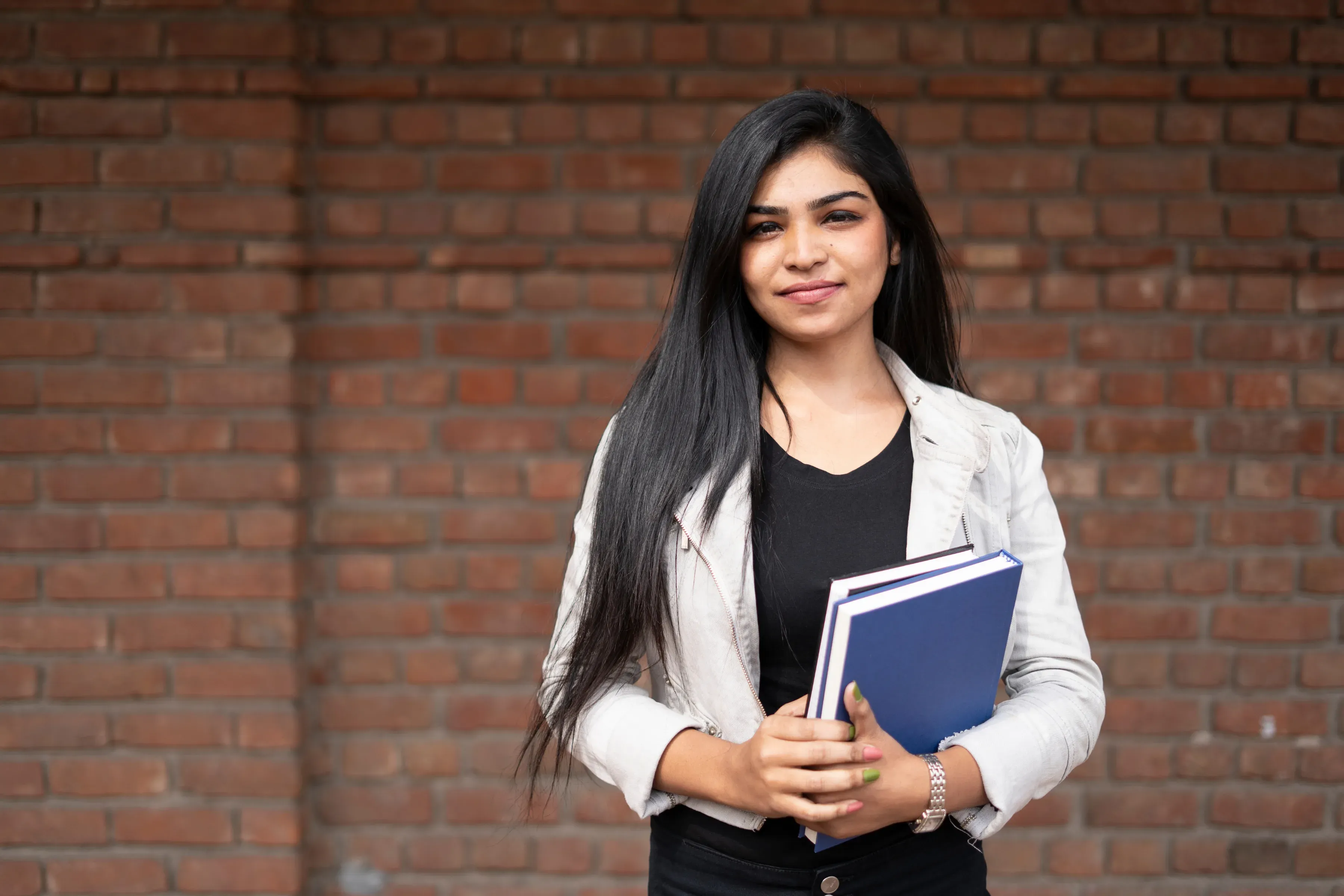 A student holding course books in front of a brick wall.